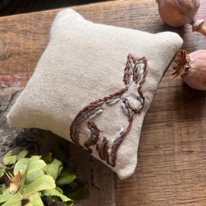 May include: A small, square, beige linen pincushion featuring an embroidered outline of a rabbit. The embroidery uses dark brown and light grey thread to define the shape of the sitting hare. The cushion is filled to create a soft, plump appearance.