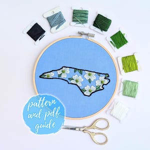 May include: A wooden embroidery hoop containing light blue fabric with an embroidered outline of North Carolina filled with white dogwood flowers. Surrounding the hoop are eight spools of embroidery thread in various shades of green and white, along with gold-toned metal scissors and a blue circular graphic with the text pattern and pdf guide.