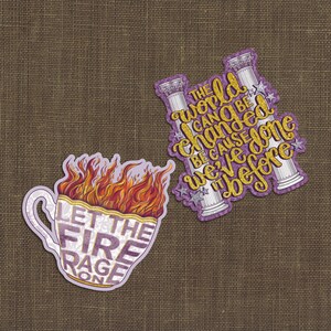 May include: Two die-cut stickers. One is a teacup with orange and red flames and the text "LET THE FIRE RAGE ON". The other features two white classical columns with gold glitter text reading "THE WORLD CAN BE CHANGED BECAUSE WE'VE DONE IT BEFORE".