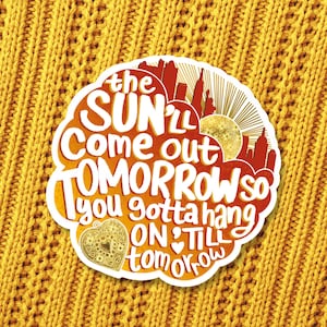 May include: A die-cut sticker with a gradient orange and red background featuring a silhouette of a city skyline and a sun. The text reads: the SUN'LL Come out TOMORROW so you gotta hang ON 'TILL tomorrow. A gold heart icon is at the bottom left.