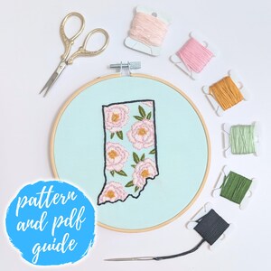 Può includere: Un telaio da ricamo con un tessuto azzurro chiaro con un contorno ricamato dello stato dell'Indiana, riempito con disegni floreali rosa. Sono presenti anche forbici dorate, rocchetti di filo di vari colori e un ago. Il testo sull'immagine recita "pattern and pdf guide."