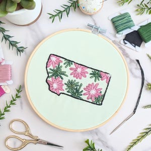 May include: A close-up of a hand-stitched embroidery hoop featuring a floral design of pink flowers and green leaves in the shape of the state of Montana. The hoop is on a white marble surface with embroidery floss and scissors.