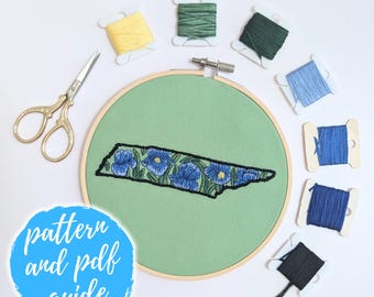 May include: Embroidery hoop with a green fabric background and a black outline of the state of Tennessee filled with blue flowers. The hoop is surrounded by spools of embroidery floss in various colors, including green, yellow, blue, and black.