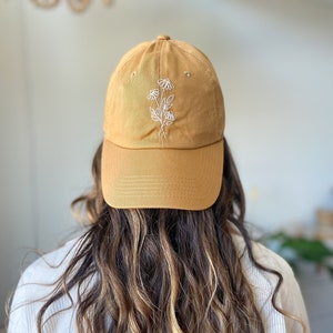 Puede incluir: Una gorra de béisbol de color amarillo mostaza con un diseño floral bordado en blanco.