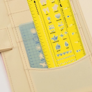 May include: A yellow and blue plastic ruler with a variety of shapes and symbols for drawing and journaling. The ruler is in a beige pocket of a planner.