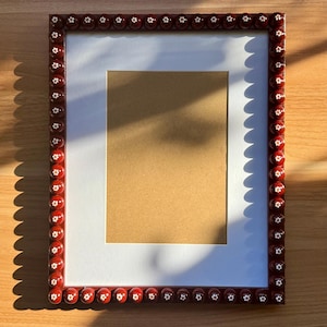 May include: A rectangular picture frame with a dark red border. The border is decorated with small, white flower-shaped accents. The frame has a white inner border and a tan backing, perfect for displaying photos or artwork.