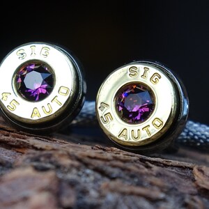 May include: A pair of cufflinks made from .45 Auto shell casings with a purple gemstone set in the center of each casing. The casings are engraved with "SIG AUTO".