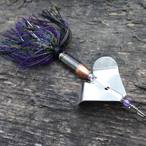May include: A fishing lure with a silver body, copper-colored tip, and a purple and black skirt. The lure has a heart-shaped blade and a small purple bead. The lure is designed for catching fish.