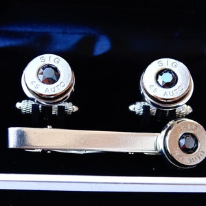 May include: A set of silver cufflinks and a tie clip presented in a black box. The cufflinks and tie clip feature a bullet design with a black gemstone center and the text "SIG 45 AUTO".