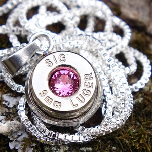 May include: A silver necklace with a 9mm bullet casing pendant featuring a pink gemstone and the inscription "SIG LUGER".
