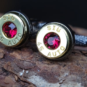 May include: A pair of cufflinks with a unique design. Each cufflink features a brass-colored casing with the text "SIG 45 AUTO" and a vibrant red gemstone center. The cufflinks have a dark, textured metal backing.