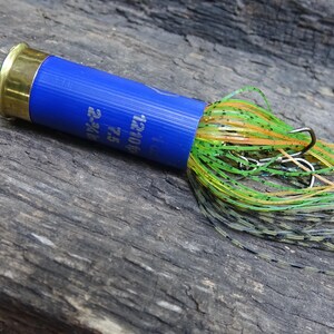 May include: A blue and gold fishing lure with a hook and colorful skirt. The lure has a blue body with white text and a gold-colored end. The skirt is a mix of green, orange, and black strands.