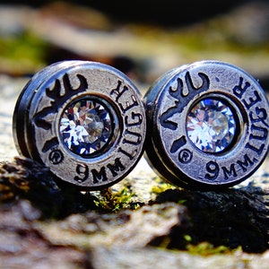 May include: A pair of silver bullet earrings with clear rhinestones. The earrings are engraved with "9MM LUGER" and a deer head design.