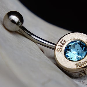 May include: A silver belly button ring with a blue gemstone set in a silver bullet charm. The charm is engraved with "SIG 10mm AUTO".