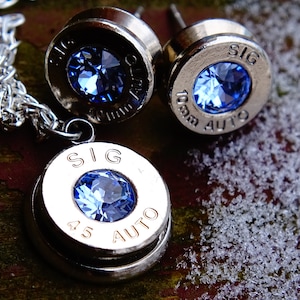 May include: A silver necklace and earrings set featuring blue crystal accents set in bullet casings. The necklace pendant reads "SIG 45 AUTO" and the earrings read "SIG 9mm AUTO".