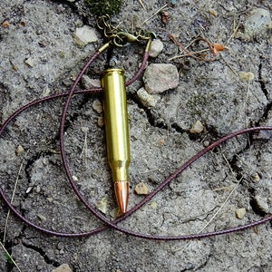 May include: A brass bullet pendant with a copper tip, suspended on a brown leather cord. The necklace is displayed on a cracked, earthy surface, showcasing a rugged, industrial aesthetic. The bullet casing is approximately 2 inches long.