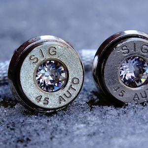 May include: A pair of silver cufflinks made from .45 caliber bullet casings with a clear rhinestone embedded in each casing. The casings are engraved with "SIG 45 AUTO".