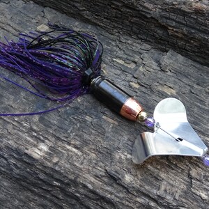 May include: A fishing lure with a black body, copper accents, and a silver blade. The lure features a purple and black skirt. The lure is designed for catching fish and is made of metal and plastic.