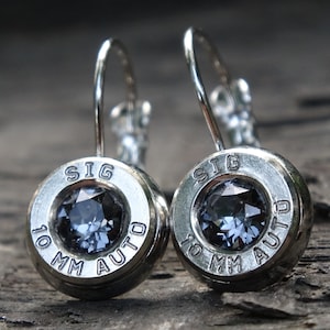 May include: A pair of silver bullet earrings with a dark blue gemstone in the center. The bullet casings are engraved with "SIG 10 MM AUTO".