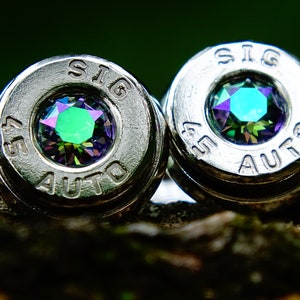 May include: A pair of silver bullet earrings with green and purple iridescent gemstones set in the center of each bullet. The bullets are engraved with "45 AUTO" and "SIG".