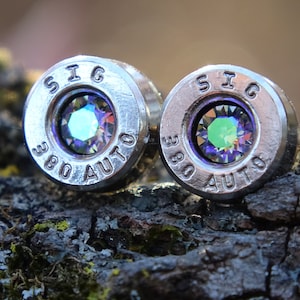 May include: A pair of silver bullet earrings with iridescent gemstones set in the center. The bullets are engraved with "SIG 380 AUTO".