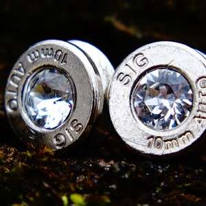 May include: A pair of silver bullet earrings with clear rhinestones. The earrings are engraved with "SIG" and "7.62 mm AUTO".