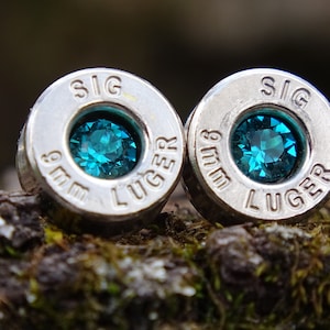 May include: A pair of silver bullet earrings with turquoise gemstones. The bullets are engraved with "9mm SIG LUGER".