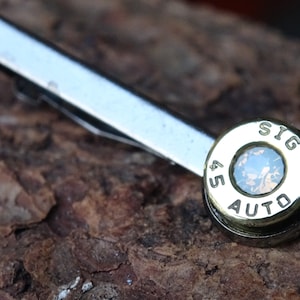 May include: A silver tie clip with a gold-colored .45 SIG AUTO bullet casing as the decorative element. The bullet casing has a clear stone in the center.
