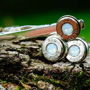 May include: Silver tie clip with three bullet casings, each with a white opal stone and the inscription "SIG 45 AUTO".