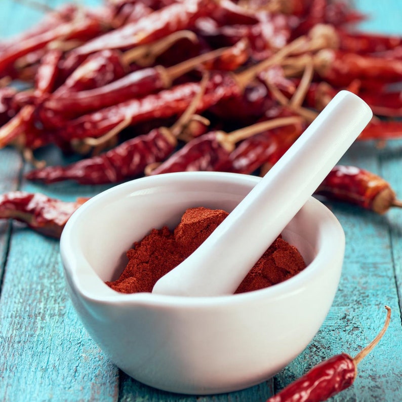 May include: A white ceramic mortar and pestle filled with red powder, alongside a pile of dried red chili peppers. The mortar and pestle sit on a weathered, light blue wooden surface. The image highlights the preparation of spices.
