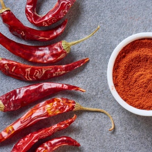 May include: Dried red chili peppers and a white bowl filled with red chili powder on a gray surface. The peppers are long and slender with green stems. The powder has a fine texture.