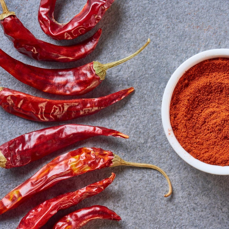 May include: Dried red chili peppers and a white bowl filled with red chili powder on a gray surface. The peppers are long and slender with green stems. The powder has a fine texture.
