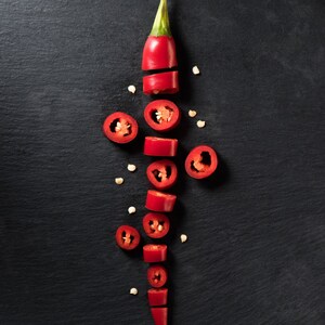 May include: A top-down shot of a sliced red chili pepper on a black surface. The pepper is cut into several pieces, revealing the seeds inside. The pieces are arranged vertically, with a few seeds scattered around.