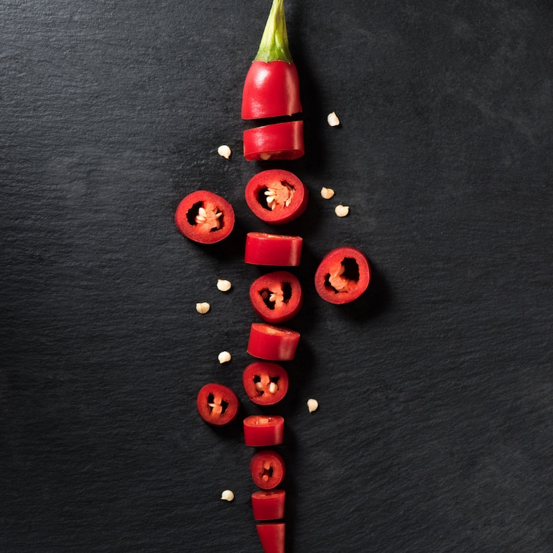May include: A top-down shot of a sliced red chili pepper on a black surface. The pepper is cut into several pieces, revealing the seeds inside. The pieces are arranged vertically, with a few seeds scattered around.