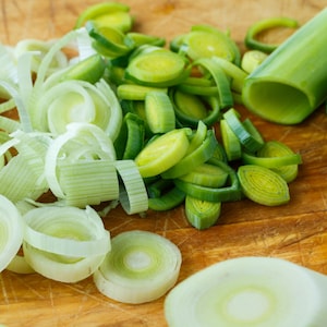 May include: Close-up of a wooden cutting board with sliced leeks. The leeks are cut into thin rounds and some are sliced lengthwise. The leeks are a light green color.
