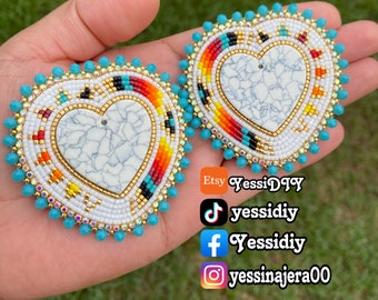 May include: A pair of heart-shaped earrings with turquoise beads and a white heart-shaped stone in the center. The earrings are surrounded by a beaded design with red, orange, yellow, and black beads.