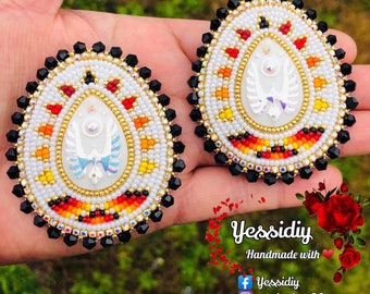 May include: A pair of white beaded earrings with a red, orange, and yellow geometric pattern. The earrings have a white iridescent center with a silver design. The earrings are handmade and are shown on a hand.