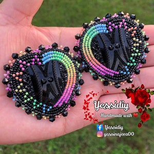 May include: Heart-shaped beaded earrings with a black center and a rainbow gradient border. The earrings are handmade with black beads and a black center design. The earrings are held in a hand.