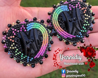 Può includere: Orecchini a forma di cuore fatti a mano con perline. Gli orecchini hanno un centro nero e un bordo sfumato arcobaleno. Gli orecchini sono tenuti in una mano.