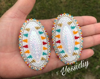 May include: A pair of oval-shaped earrings with white, red, orange, turquoise, and gold beads. The earrings have a white iridescent center with a snowflake design.
