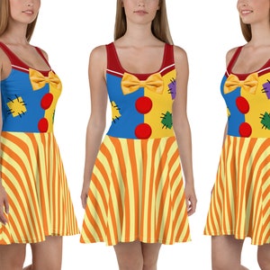 Puede incluir: Vestido sin mangas con temática de payaso, con cuello y tirantes rojos. El corpiño es azul y amarillo con un lazo amarillo, círculos rojos y parches. La falda presenta rayas verticales naranjas y amarillas.