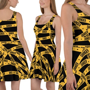Puede incluir: Un vestido skater negro con un patrón de cinta de advertencia amarilla y negra. El vestido tiene la palabra "DANGER" impresa repetidamente en la cinta.