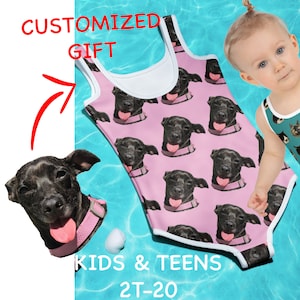 May include: Pink swimsuit with a repeating pattern of a black dog's face. The swimsuit has white trim around the neck and armholes. The text "KIDS & TEENS 2T-20" is printed on the bottom of the swimsuit.