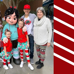 May include: A family of five pose for a photo with a character from the film Wreck-It Ralph. The character is wearing a blue and white outfit and has black hair. The family are wearing festive holiday attire, including red and white striped leggings, a gingerbread house sweater, and a red and white striped shirt. The character is holding a child in a red shirt and a green and white skirt.