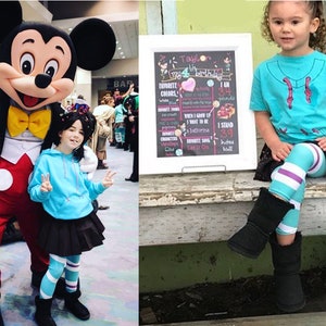 May include: A girl dressed as Vanellope von Schweetz from Wreck-It Ralph stands next to Mickey Mouse. The girl is wearing a blue hoodie, a black skirt, and striped leggings. She is holding up two fingers in a peace sign. Mickey Mouse is wearing his signature red shorts and yellow gloves. The girl is smiling and looking at the camera. The background is a convention hall with people walking around.
