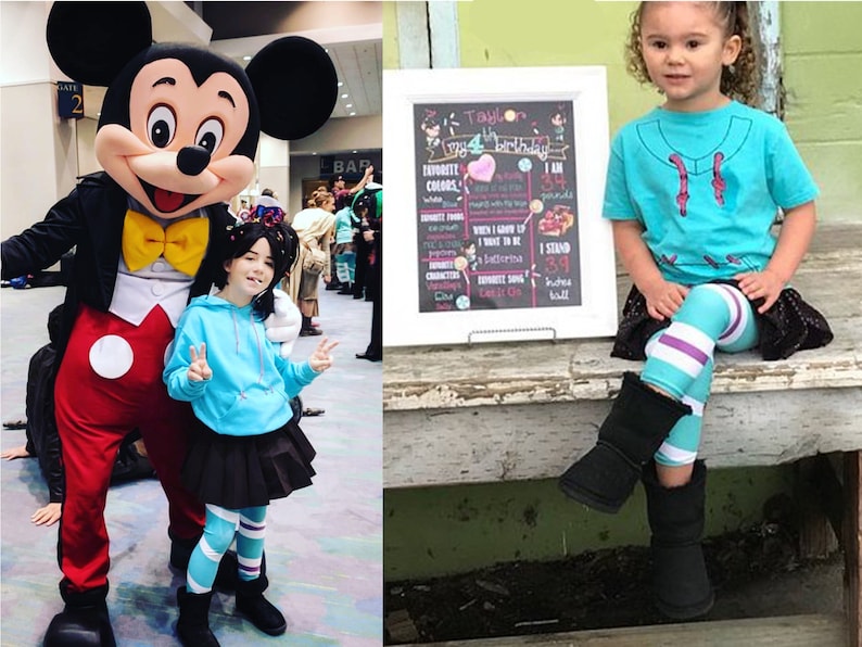 May include: A girl dressed as Vanellope von Schweetz from Wreck-It Ralph stands next to Mickey Mouse. The girl is wearing a blue hoodie, a black skirt, and striped leggings. She is holding up two fingers in a peace sign. Mickey Mouse is wearing his signature red shorts and yellow gloves. The girl is smiling and looking at the camera. The background is a convention hall with people walking around.