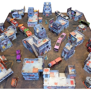 Op de afbeelding: Een tabletop-spelscène met miniatuurgebouwen in verschillende kleuren, waaronder grijs, rood en roze. De gedetailleerde structuren zijn gerangschikt op een bruin oppervlak, waardoor een stadsbeeld ontstaat.