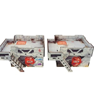 Op de afbeelding: Twee miniatuur, grijze, lasergesneden gebouwen met een futuristisch, industrieel ontwerp. Elk gebouw heeft een dak, een trap en graffiti-kunst. De gebouwen hebben rode en oranje accenten en de tekst "ACCESS HATCH" is zichtbaar op het dak.