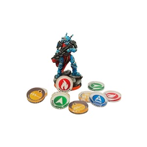 Puede incluir: Una figura en miniatura azul y roja de un marine espacial sosteniendo un arma, de pie sobre una base con un s&iacute;mbolo de llama. La figura est&aacute; rodeada de ocho fichas circulares con diferentes s&iacute;mbolos, incluyendo una llama, una estrella, un rayo y un coraz&oacute;n.