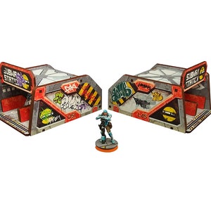Puede incluir: Dos edificios modelo de cartón pintados de gris y rojo con grafitis y el texto "Subway Station" en el lateral. Una figura en miniatura azul y gris se encuentra frente a los edificios.